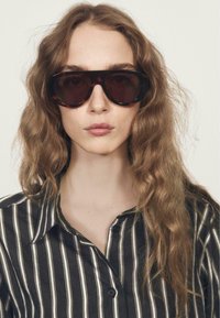 Przesadnie duże czarne okulary przeciwsłoneczne z ciemnymi szkłami, na tle modelki noszącej czarną i białą w paski koszulę z kołnierzykiem. Widoczne fale włosów.