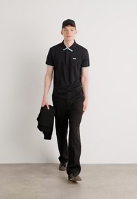 Czarna koszulka polo z kontrastującymi akcentami kołnierzyka, zestawiona z czarnymi spodniami. Model trzyma złożoną parę spodni, nosząc czarną czapkę.