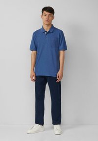 Niebieska koszulka polo z fakturowanej tkaniny, z kołnierzykiem i kieszonką na piersi, w połączeniu z ciemnoniebieskimi spodniami i białymi trampkami.