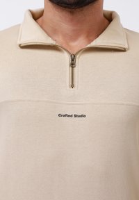 Sweatshirt beige zippé avec un tissu texturé, présentant un col montant et un petit logo "Crafted Studio" sur le devant.