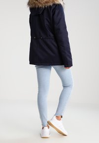 Osoba oblečená v námořnické parce s kožíškem na kapuci, světle modrých úzkých džínech a bílých teniskách, stojící na jedné noze s zády k fotoaparátu.