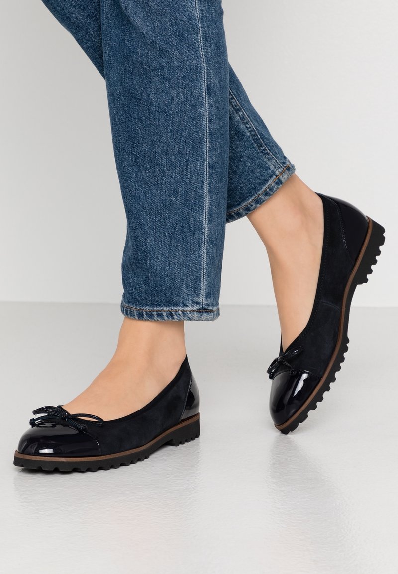 Stopy w czarnych, lakierowanych i zamszowych balerinach z kokardkami, w połączeniu z niebieskimi dżinsami na jasnej podłodze.