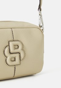 Beige skinnveske med kryssbærerem, med en teksturert overflate, som har en fremtredende preget logo, glidelås lukking, og en avtagbar stropp.