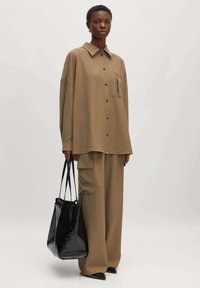 Braunes, oversized Hemd mit einer Tasche, kombiniert mit lockeren, passenden Hosen. Eine schwarze, glänzende Schultertasche und spitze schwarze Schuhe runden das Outfit ab.