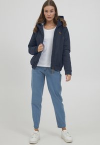 Giacca blu navy con zip frontale, cappuccio e polsini elastici. Materiale leggero, con un discreto dettaglio del logo. Indossata sopra una camicia bianca e jeans blu.