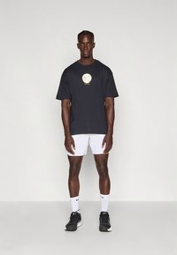 Svart bomulls-T-skjorte med en gyllen Nike-logo, kombinert med hvite shorts. Modellen har på seg svarte joggesko og hvite sokker, og står foran en nøytral bakgrunn.