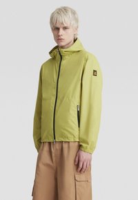 Limegrüner Reißverschlussjacke mit Kapuze, schwarzem Reißverschluss, Seitentaschen und elastischen Bündchen. Lässiges Design kombiniert mit sandfarbenen Cargo-Shorts.
