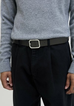 Ceinture en cuir noir avec une boucle en argent rectangulaire, portée sur un pantalon noir et sous un pull en laine gris.