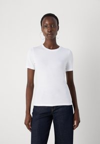 Femme aux cheveux courts portant de grandes boucles d'oreilles anneaux, un t-shirt blanc uni à manches courtes et un jean bleu foncé, debout devant un fond blanc.