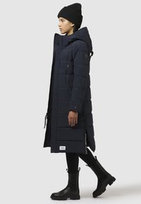 Lungo cappotto imbottito blu navy con cappuccio, tasche con zip e polsini a coste. Abbinato a stivali neri alla caviglia e un berretto di lana.