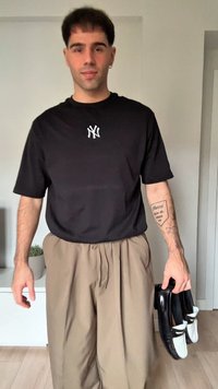 Jovem usando uma t-shirt preta dos New York Yankees e calças bege segurando sapatos mocassins preto e branco em ambientes fechados, mostrando uma tatuagem no antebraço.