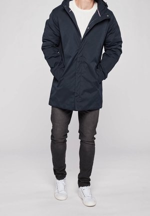 Mies, jolla on päällään laivastonsininen hupullinen talvitakki, valkoinen villapaita, tummat farkut ja valkoiset lenkkarit, seisoo kädet takin taskuissa.