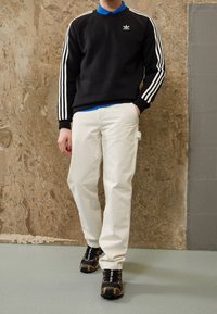 Man som bär en svart Adidas-tröja med vita ränder, en blå skjorta under, krämfärgade byxor och svarta sneakers med orange detaljer.