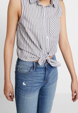 Szara i biała koszula bez rękawów z pionowymi paskami, zapinana na guziki i zawiązana w talii, noszona z niebieskimi postrzępionymi dżinsami na stojącej osobie.