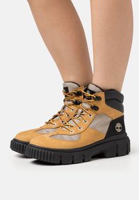 Bottes de randonnée en daim marron avec accents noirs, détails en mesh et lacets jaunes. Dotées d'une semelle épaisse noire texturée avec un logo sur le côté.
