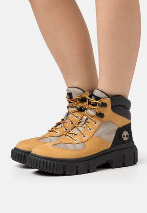 Bottes de randonnée en daim marron avec accents noirs, détails en mesh et lacets jaunes. Dotées d'une semelle épaisse noire texturée avec un logo sur le côté.