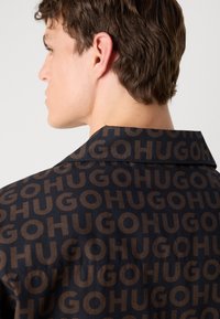 Camisa negra con un gran logotipo repetido "HUGO" en marrón, con una textura lisa y un diseño de cuello clasico.