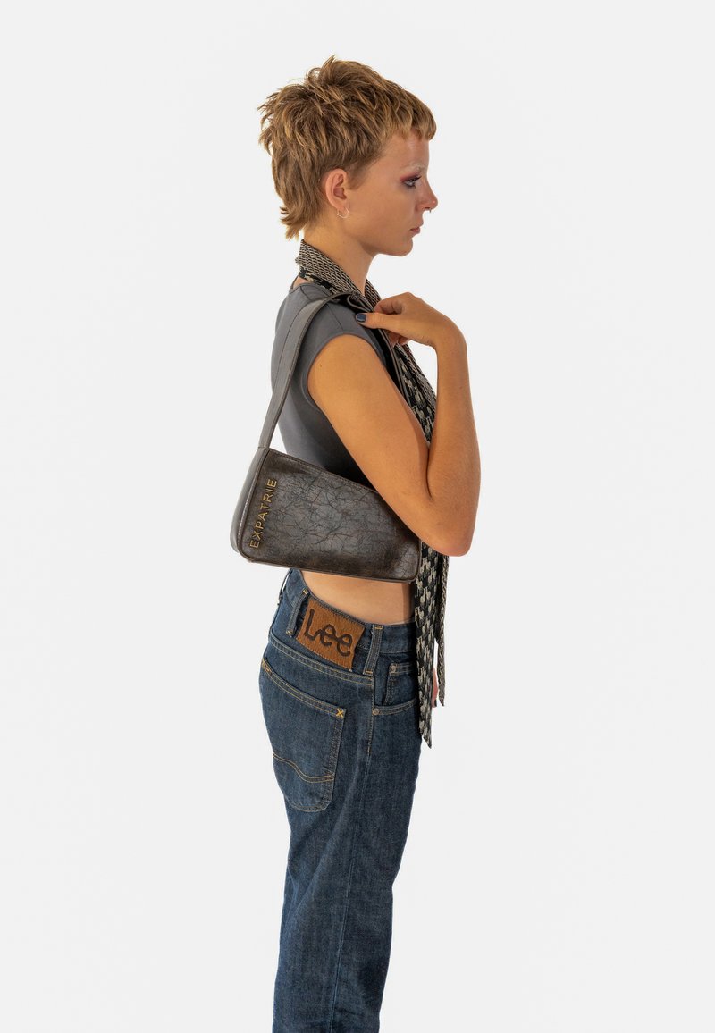 Borse in pelle grigia a forma triangolare, con il nome del marchio "EXPATRIÉ" in rilievo e in oro, abbinata a una sciarpa a motivi bianco e nero e jeans blu.