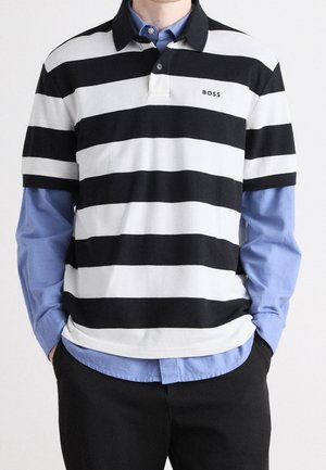 Person iført en sort/hvid horisontalt striped kortærmet polo over en blå langærmet skjorte med sorte bukser, hænderne i lommerne.