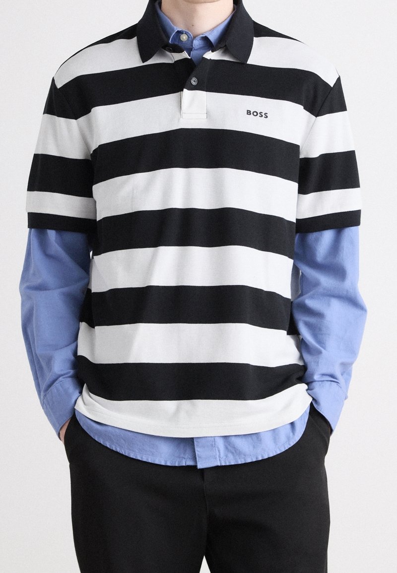 Personne portant un polo à manches courtes à rayures horizontales noires et blanches sur une chemise bleue à manches longues avec un pantalon noir, les mains dans les poches.