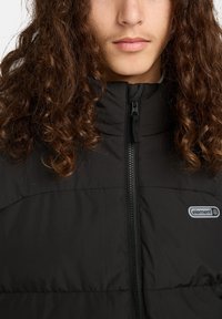 Schwarze gepolsterte Jacke mit hohem Kragen, Frontreißverschluss und einem kleinen Logo-Patch auf der linken Brust. Der Stoff wirkt glatt und isoliert.