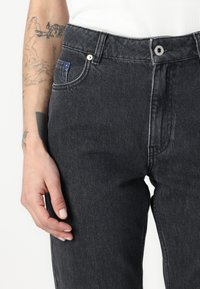 Melnas džinsu bikses ir ar augstu jostasvietu, priekšējiem kabatām un minimālu šuvju skaitu. Uz priekšējās kabatas ir redzams neliels zils akcenti.