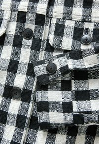 Czarno-biała tkanina w kratkę o teksturowanym splocie. Z czarnymi guzikami i podwójnym wzorem kieszeni. Zbliżenie na detale.