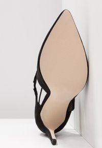 Chaussure à talon haut noire avec un bout pointu, en matière suédée, et un talon stiletto fin. La semelle est beige avec une finition texturée.