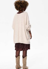 Beżowy, oversize sweter z podwiniętymi rękawami, nałożony na ciemnogranatową spódnicę z koronkowymi wykończeniami, w zestawieniu z beżowymi kowbojkami.