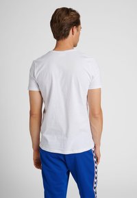 Mand med kort brunt hår iført en ensfarvet hvid t-shirt med korte ærmer og blå bukser med en sort-hvid mønstret sideliste, set bagfra.