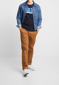 Denim jacket, navy t-shirt with blue graphic, brown trousers, and white sneakers; model stands with hands in pockets, casual appearance.