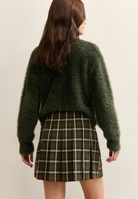 Maglione verde peloso con un design corto, indossato sopra una gonna a pieghe a quadri con accenti verde scuro, bianco e rosso.