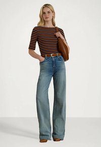 Ženska v mornarsko modri in rjavi črtasti majici, hlačah z visokim pasom in širokimi nogavicami, rjavem pasu, rjavih čevljih in z rjavo torbo na rami.