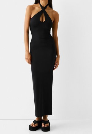 Žena v černých maxi šatech bez rukávů s halenkovým výstřihem a průstřihem vpředu, obutá do černých sandálů na platformě, na jednoduchém bílém pozadí.