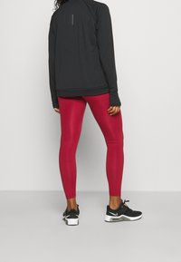 Person steht mit dem Rücken zu uns und trägt ein schwarzes Langarmshirt, leuchtend rote Leggings und schwarze sowie weiße Nike-Sneaker vor einem einfarbigen Hintergrund.