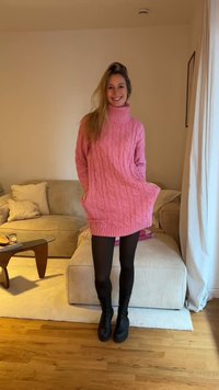 Różowa, oversize'owa sukienka swetrowa z grubym splotem, z wysokim kołnierzem i kieszeniami, zestawiona z czarnymi rajstopami i botkami; minimalistyczne, wnętrzarskie tło.