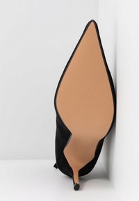 Chaussure à talon haut en daim noir avec un bout pointu et une semelle en caoutchouc beige. Le talon est fin et effilé, avec une texture lisse.