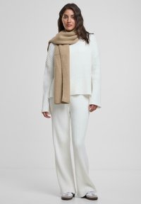 Weißer gerippter Strickpullover und passende weit geschnittene Hose, kombiniert mit einem strukturierten beigen Schal, vor einem schlichten Hintergrund stehend.