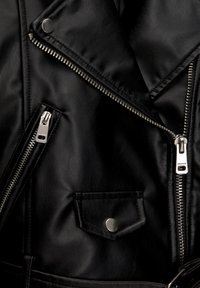 Gros plan d'une veste en cuir noire montrant des fermetures éclair argentées, un bouton-pression et une petite poche à rabat sur un matériau texturé.