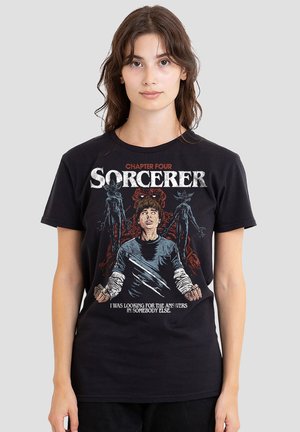 Young woman wearing black "Chapter Four Sorcerer" graphic T-shirt featuring a character with outstretched hands and two shadowy creatures behind.