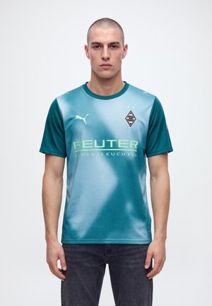 Un homme porte un maillot de football bleu canard du Borussia Mönchengladbach, orné du logo Puma et du nom d’un sponsor, sur un fond clair et uni.