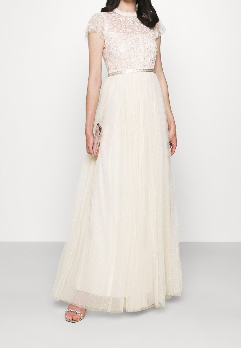 Femme portant une longue robe blanche transparente avec un corsage en dentelle florale et des manches bouffantes courtes, tenant une petite pochette et chaussant des talons ouverts.