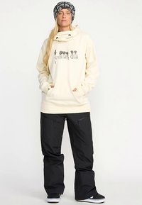 Beżowa bluza z kapturem z nadrukiem z przodu i kieszeniami kangurzymi, zestawiona z luźnymi czarnymi spodniami i czarną czapką z wzorem.