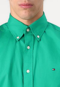Camicia in cotone verde con colletto button-down, bottoni argentati e un piccolo logo ricamato con accenti rossi, bianchi e blu.