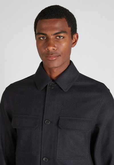 Giacca di lana scura con colletto, due tasche sul petto e bottoni neri. Il tessuto ha un aspetto liscio e strutturato.