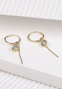 Gouden creolen met een centraal heldere kristallen hanger en een decoratieve kettingdruppel. Glad oppervlak; glanzende metalen afwerking.