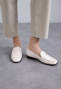 Witte leren instappers met gestikte details, lage zwarte hak en een gladde bovenkant. Draagt met lichtbeige cropped broeken op een grijze ondergrond.