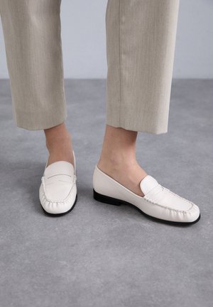 Witte leren instappers met gestikte details, lage zwarte hak en een gladde bovenkant. Draagt met lichtbeige cropped broeken op een grijze ondergrond.