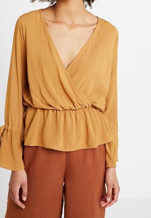 Frau trägt eine senfgelbe Wickelbluse mit V-Ausschnitt und langen Ärmeln sowie rostfarbene Hosen vor weißem Hintergrund.