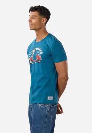 Ung mand i blå t-shirt med rød scooter grafik og tekst, stående sidelæns med hænderne bag ryggen, iført blå jeans.